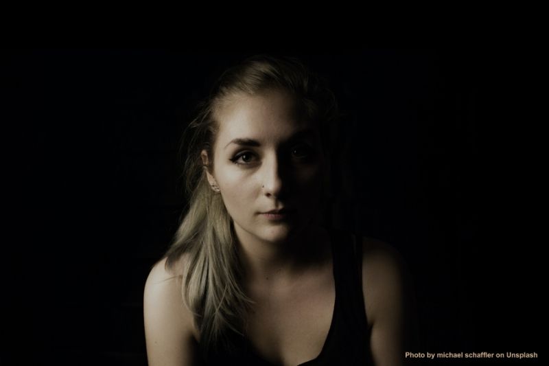 image of young white woman, likely in her 20s, with long blond hair. She is staring straight at the camera and has brown eyes. The right side of her face is in shadow. She is wearing a black tank top. The photo was taken by Michael Schaffler