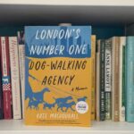a yellow and blue book titled London's Number One Dog Walking Agency: A Memoir by Kate MAcDougall sits on a white bookshelf surrounded by other memoirs