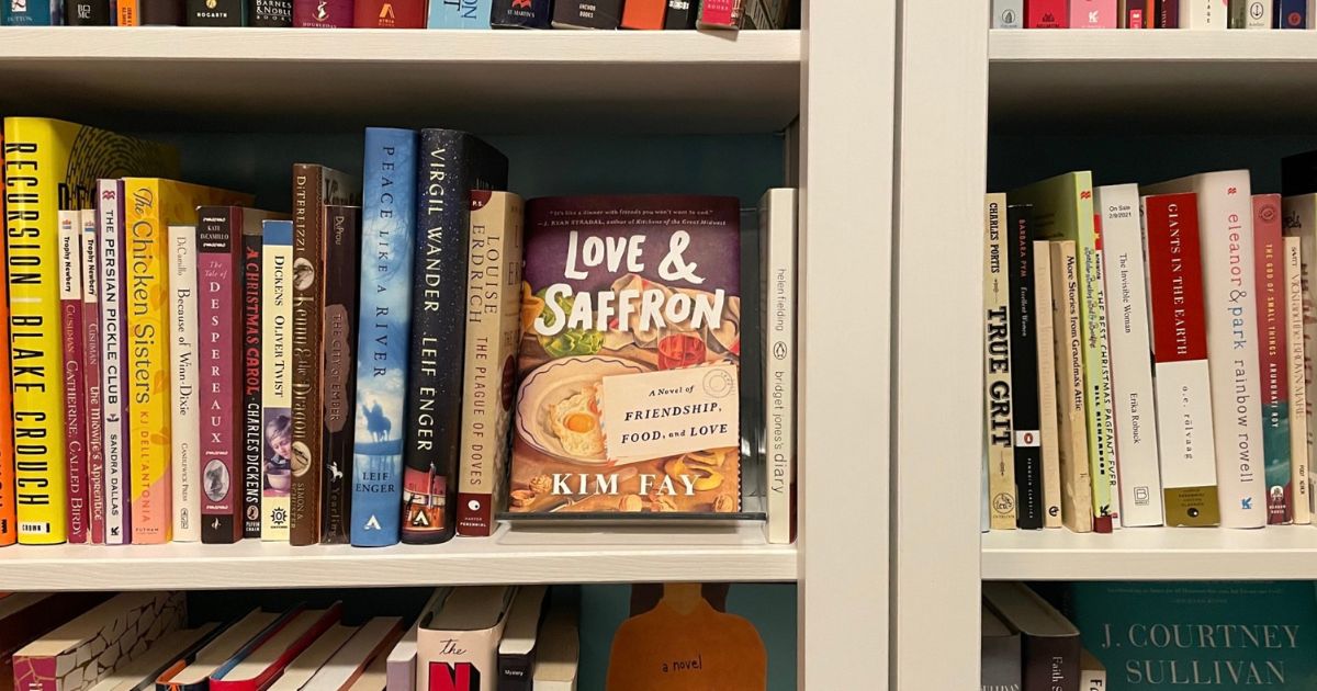 A copy of a book titled Love & Saffron by Kim Fay sits on a book shelf surrounded by many other books.