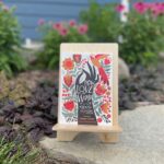 A book titled How to Love the World: Poems of Gratitude and Hope sits on a wood bookstand in a garden. The flowers and plants in the background are somewhat blurred but make for a nice backdrop for the book.