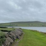 A green path with rocks on the left and the sea on the right. Green hills are in the distance and the sky is overcast with clouds