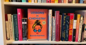A book titled Lessons in Chemistry sits on a bookshelf surrounded by other books. The book has a bright pink/orange cover with the title and author, Bonnie Games, in turquoise letters. In the center of the book is a drawn image of a woman's head, her blond hair in a bun with a pencil stuck in it. She is wearing glasses.