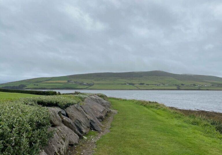 A green path with rocks on the left and the sea on the right. Green hills are in the distance and the sky is overcast with clouds